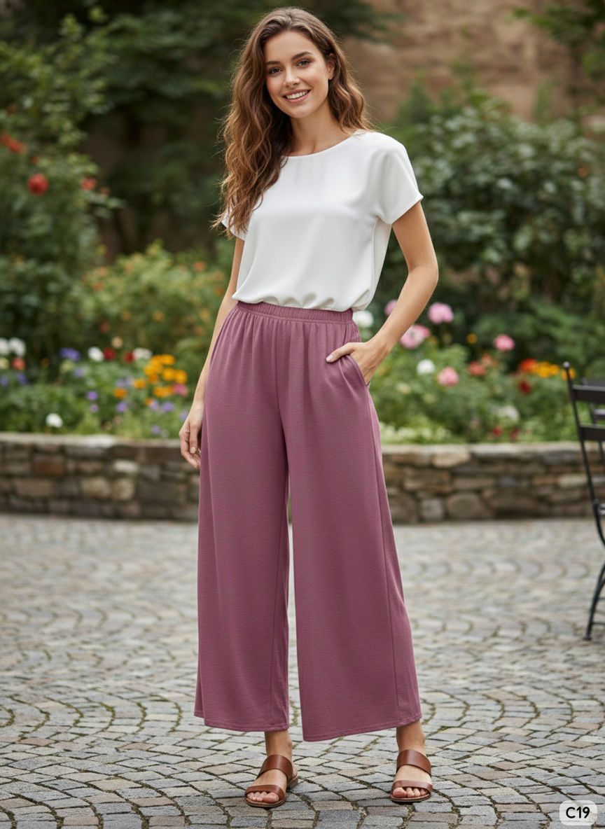 Woman wearing a white top and purple pants standing on a cobblestone path with a garden background.