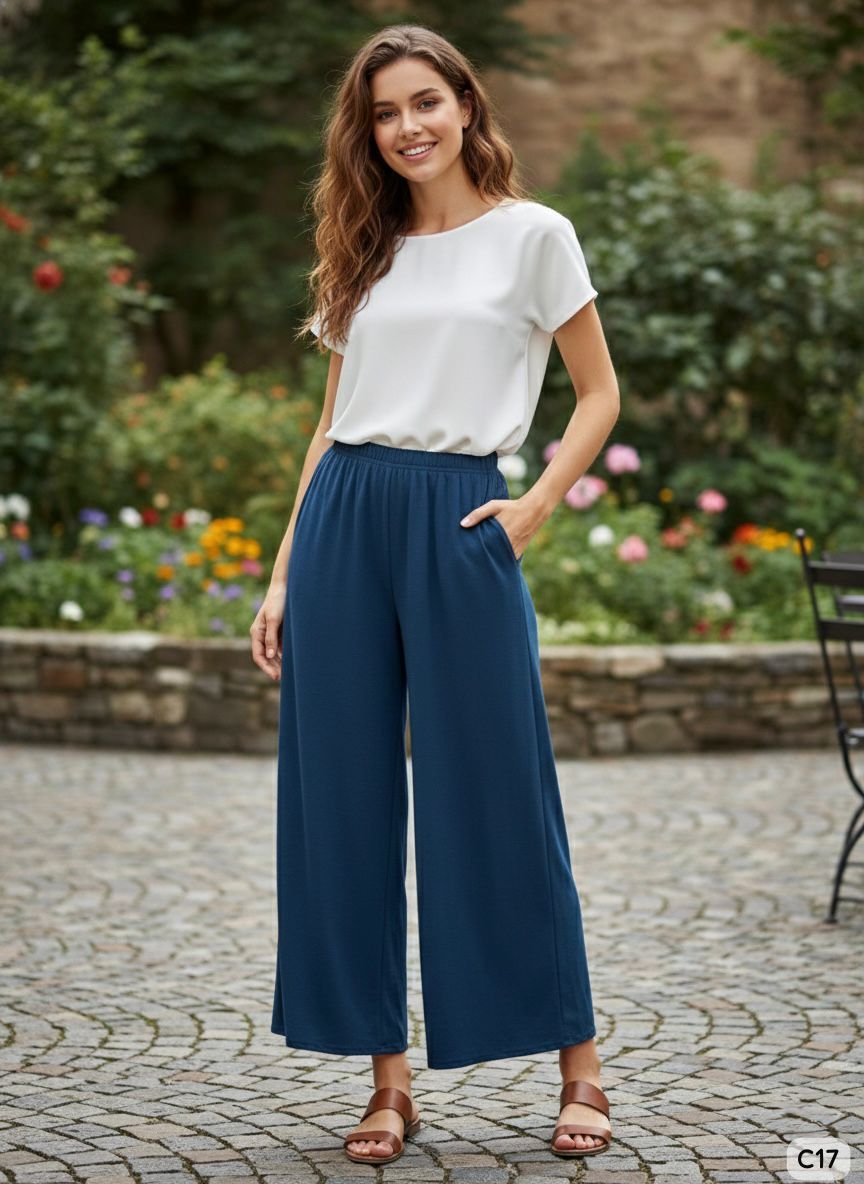 Woman wearing a white top and blue wide-leg pants standing in a garden.