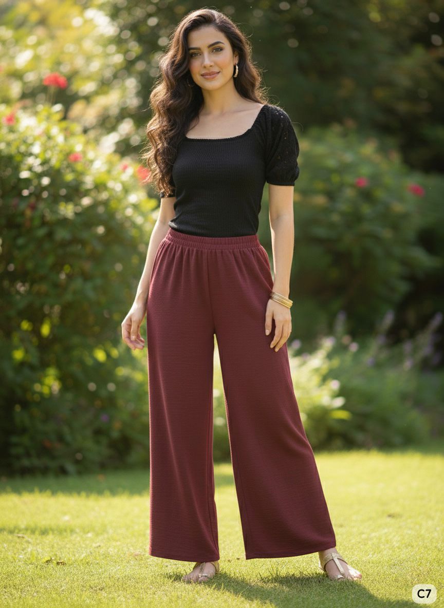 Woman wearing a black top and maroon pants standing outdoors with greenery in the background