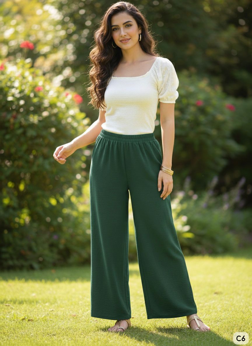 Woman wearing a white top and green pants standing outdoors with greenery in the background
