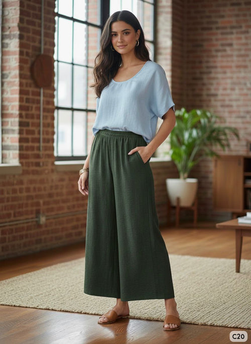 Woman wearing a light blue top and green wide-leg pants in a room with brick walls and a rug.